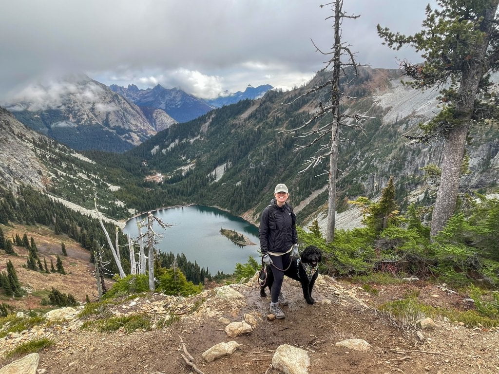 a women and a dog standing on top of a mountain over looking a lake 